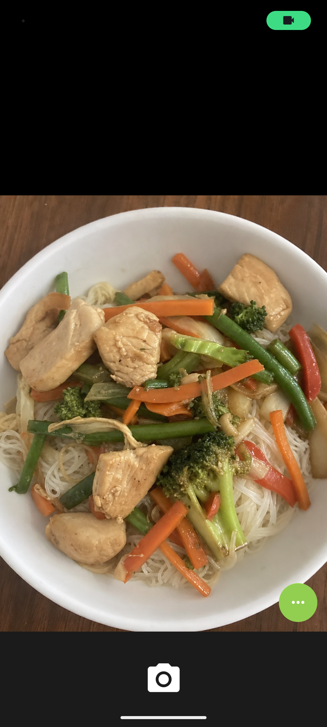 La fotocamera del telefono puntata su una ciotola di pollo, noodles di riso e verdure, pronta a fotografare il pasto